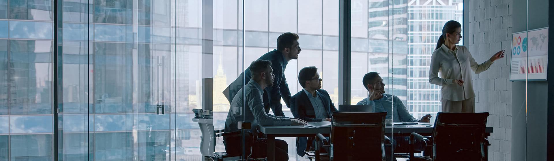 a glass-walled office with a female professional pointing to maritime shipping data and graphs on a wall monitor as three businessmen sit a table looking on. A fourth is standing also looking on.