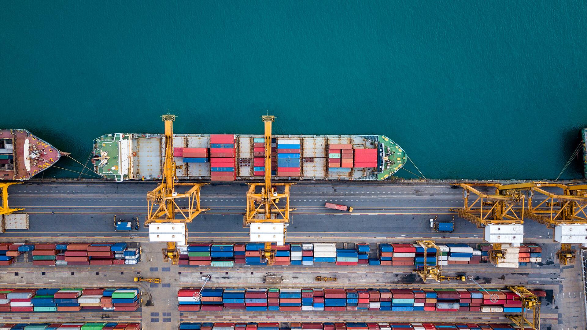 Bovenaanzicht van containerschepen in een haven. De lading van en schip wordt gelost en er hangen kranen boven. Erachter is een containerdepot te zien.